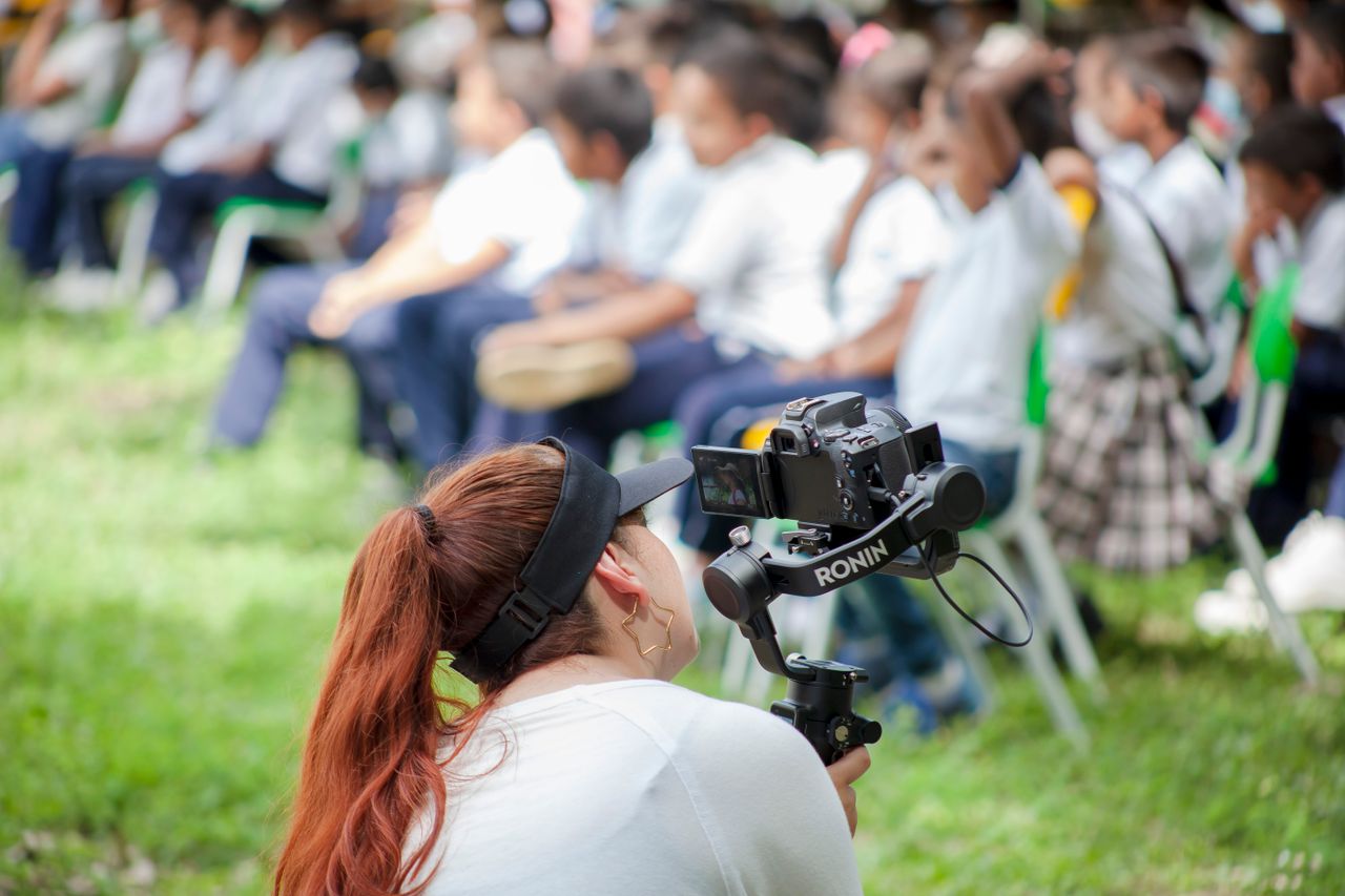 A woman videographer films a children's event outdoors in Sucre, Colombia using a stabilizer.