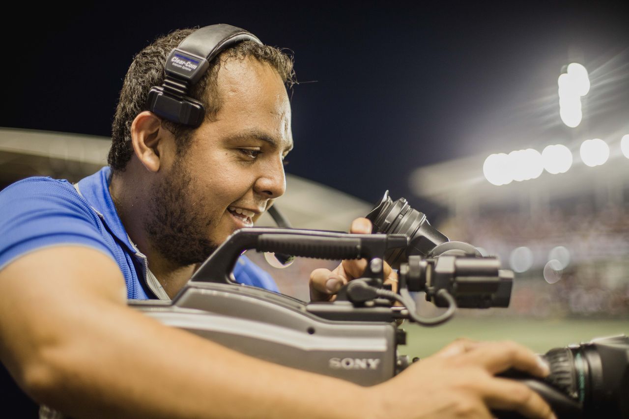 A cameraman with a headset operates a video camera outdoors at night, focusing on capturing footage.