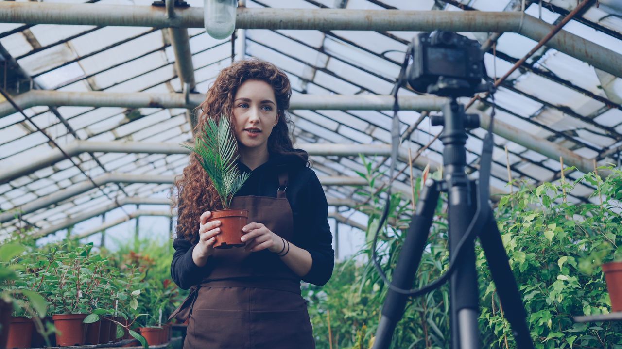 services-04 Woman in greenhouse filming a plant vlog on camera tripod.