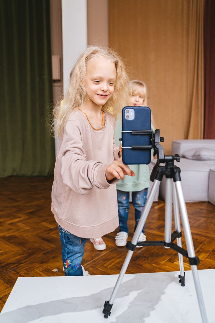 Two young girls recording with a smartphone on a tripod indoors, expressing creativity and enjoyment.